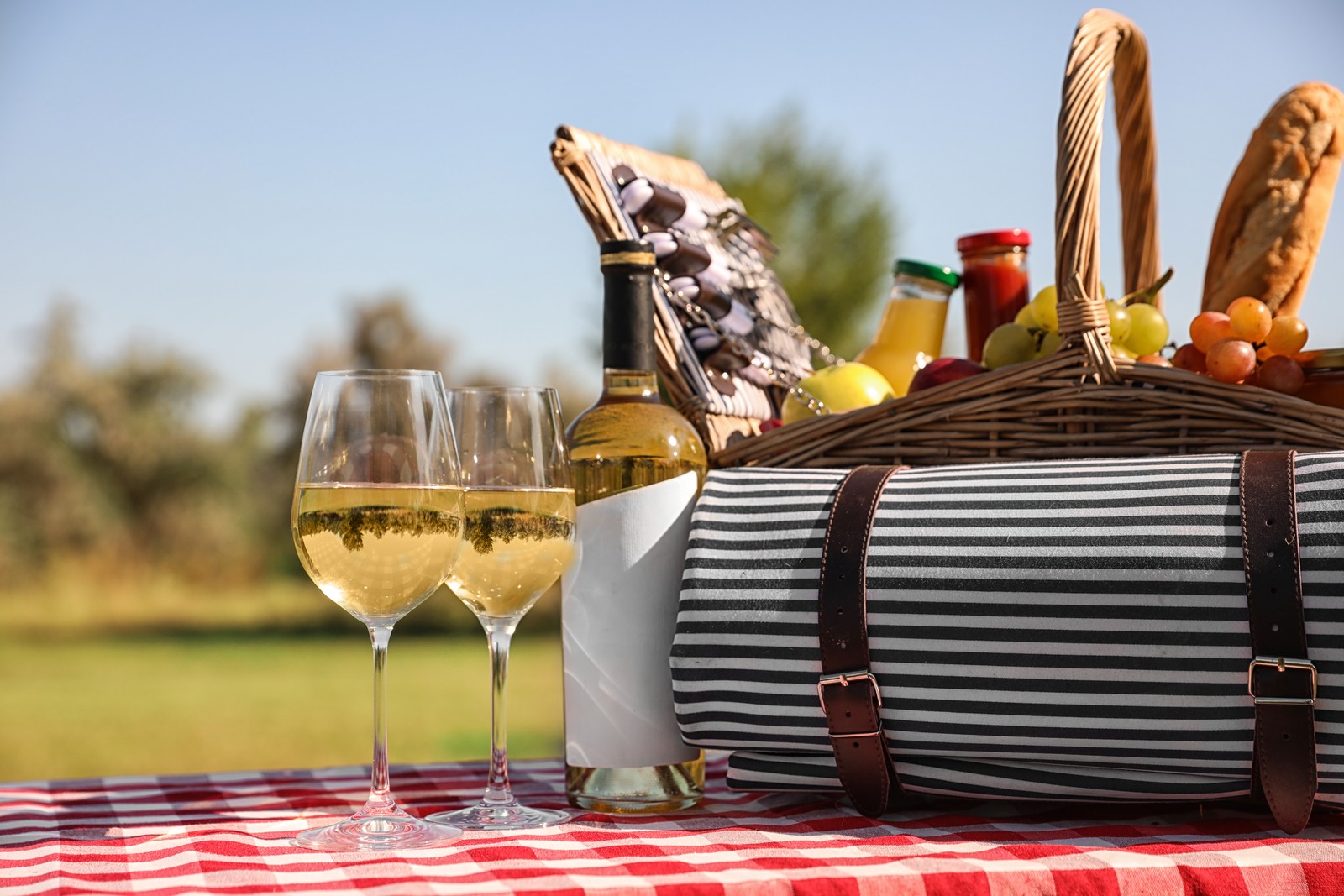 Photo of picnic basket with wine, snacks and mat on table in park