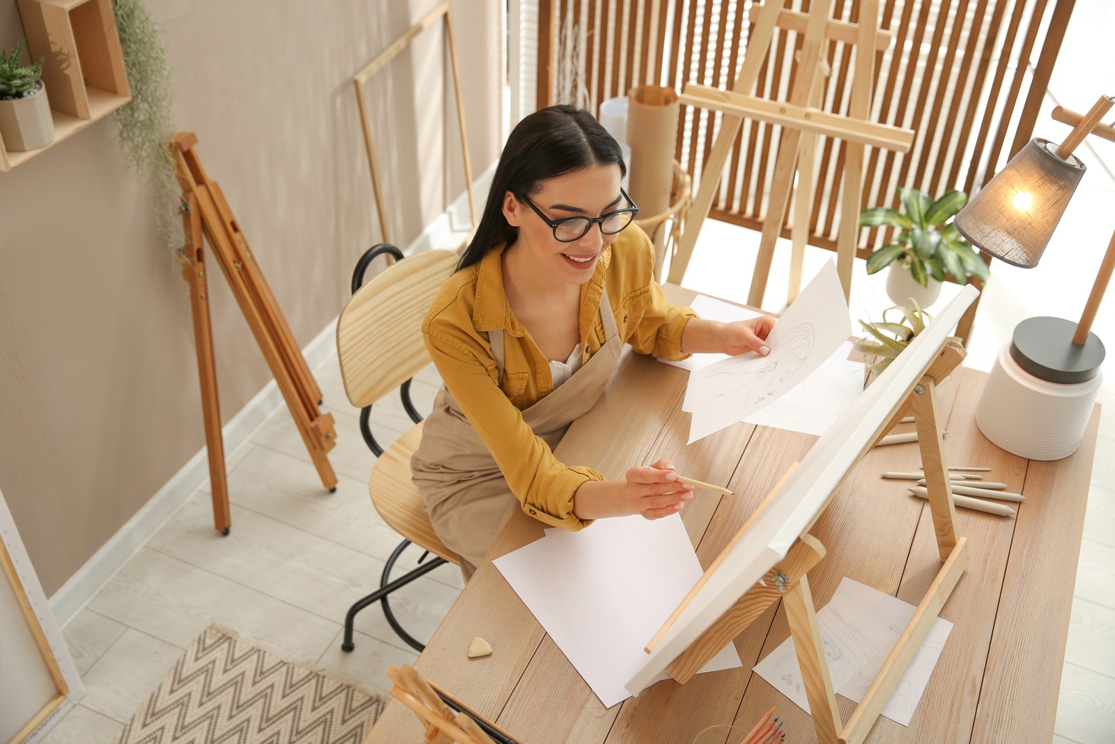 Photo of young woman drawing on easel with pencil at table indoors, above view