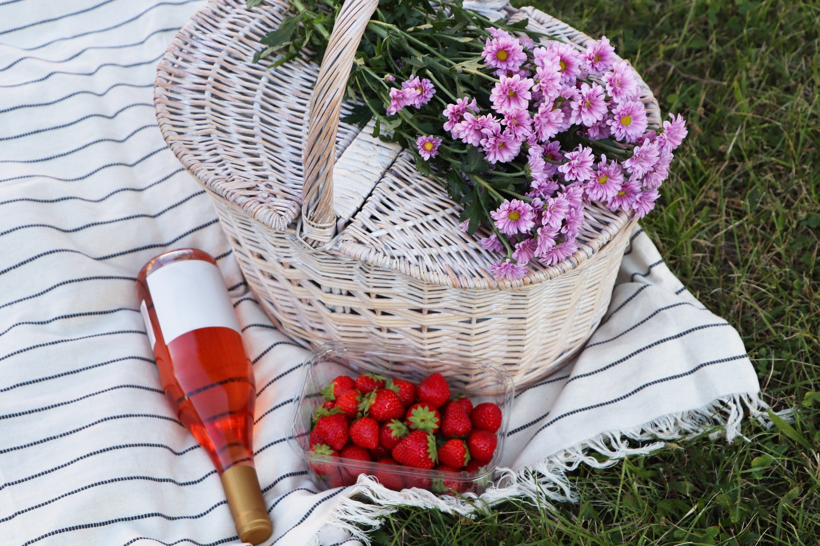 Photo of picnic basket, flowers, bottle of wine and strawberries on blanket outdoors