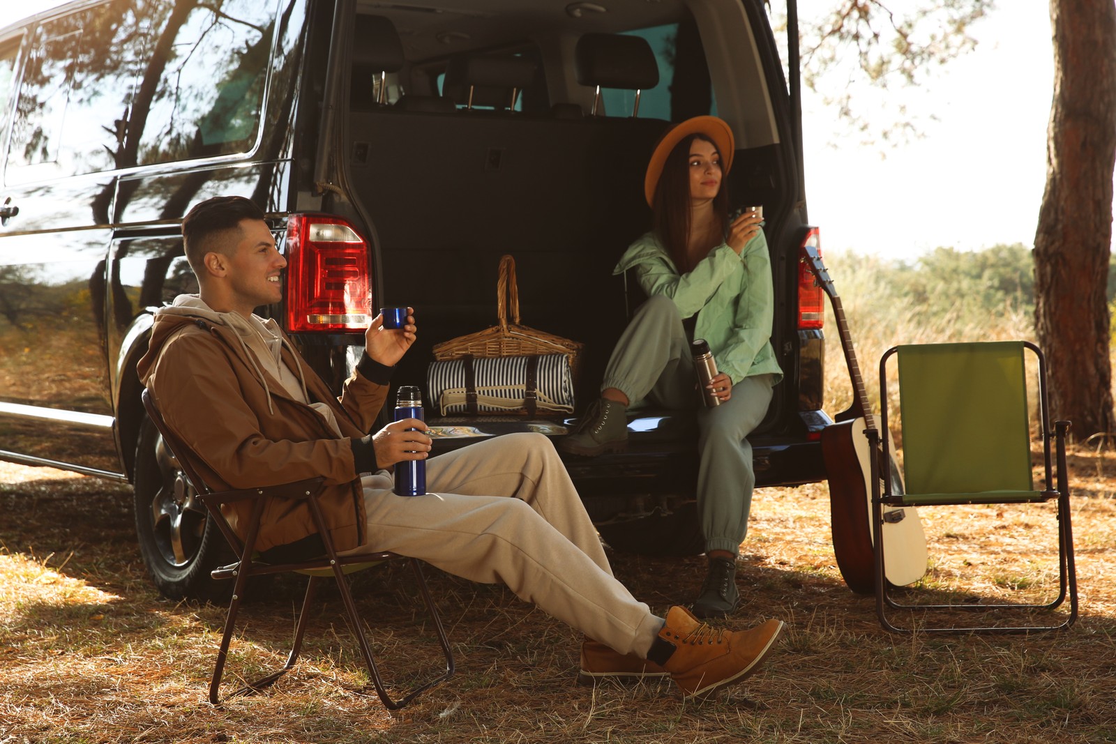 Photo of couple resting near car and enjoying hot drink at camping site