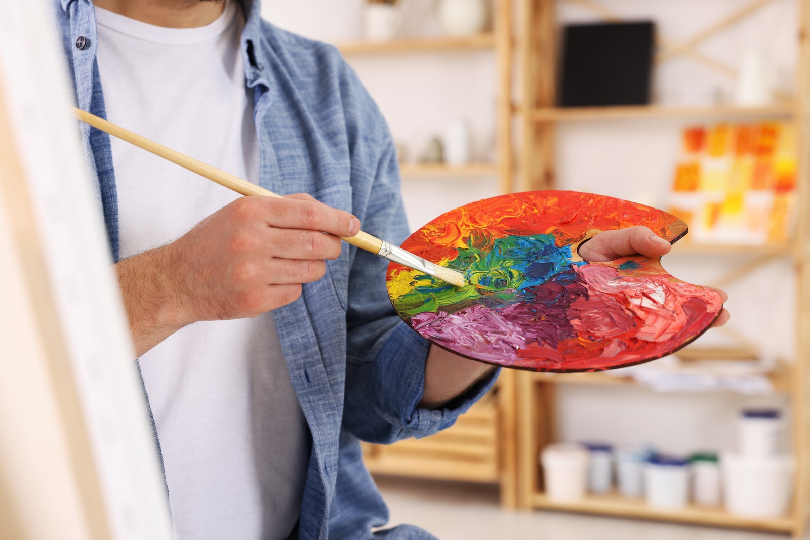 Photo of man painting in studio, closeup. Using easel to hold canvas