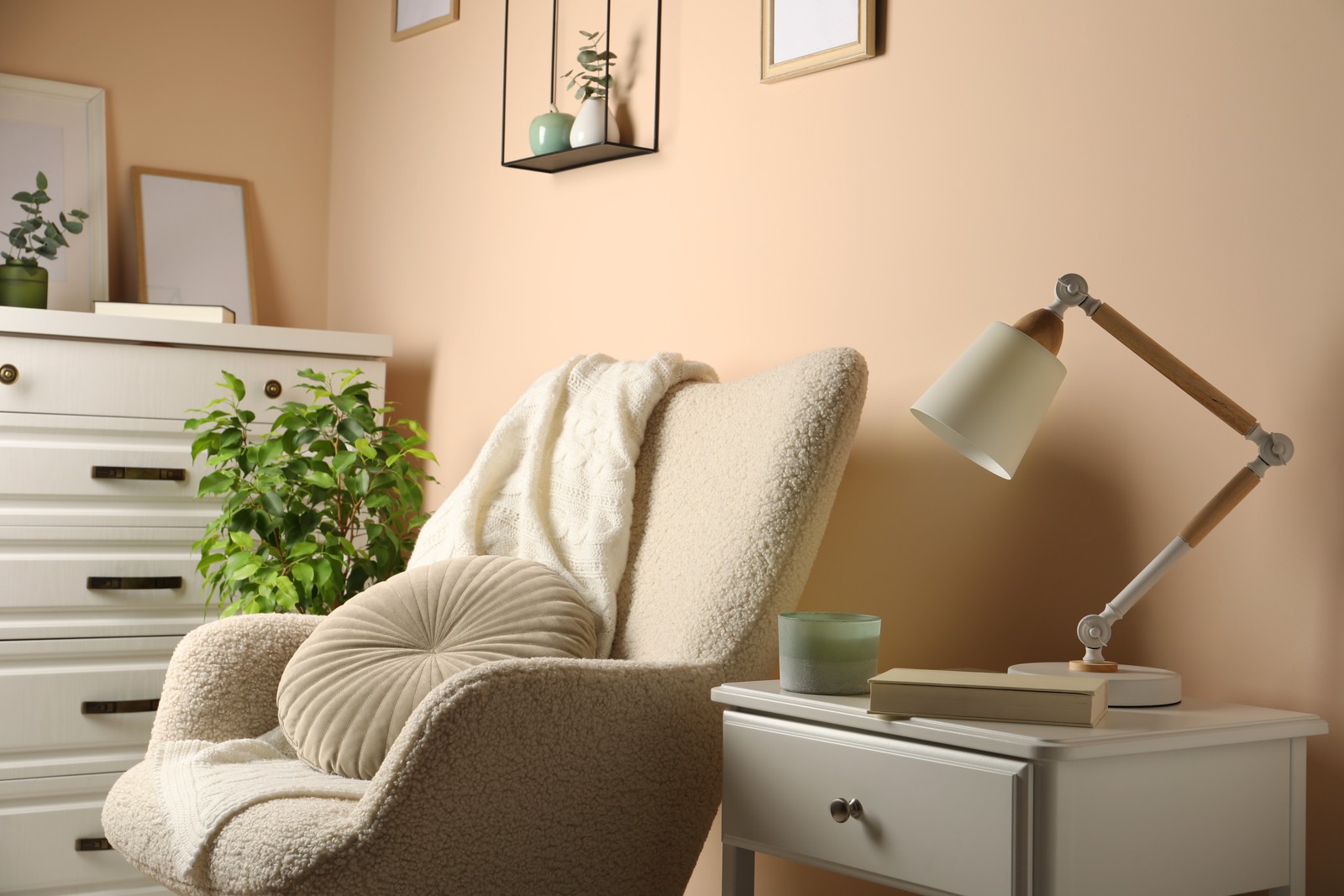 Photo of stylish modern desk lamp, book and candle on white cabinet near soft armchair indoors