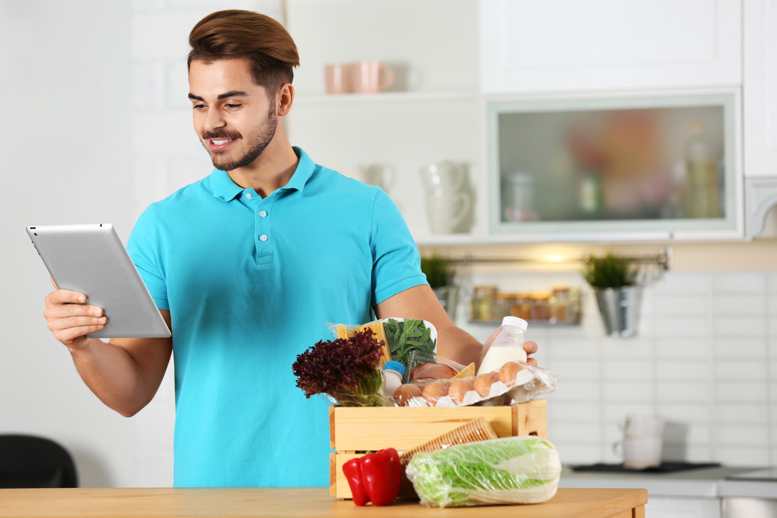 Photo of young man with tablet PC and products in kitchen. Food delivery service
