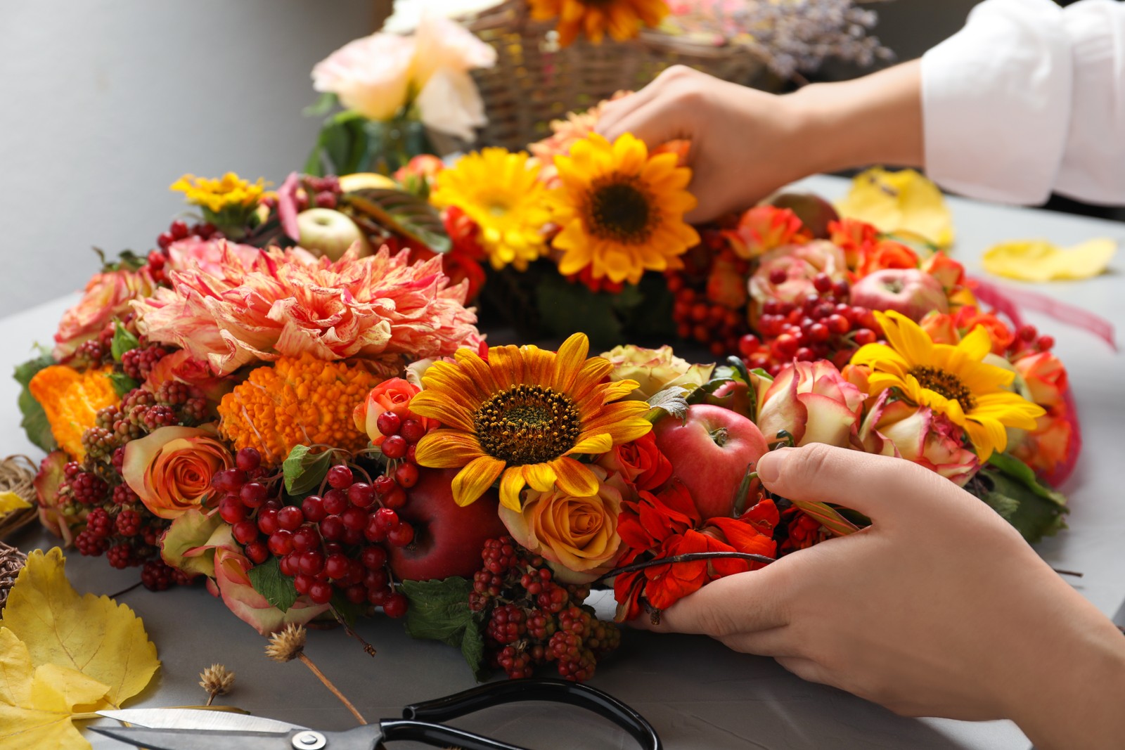 Photo of florist making beautiful autumnal wreath with flowers and fruits at light grey table, closeup