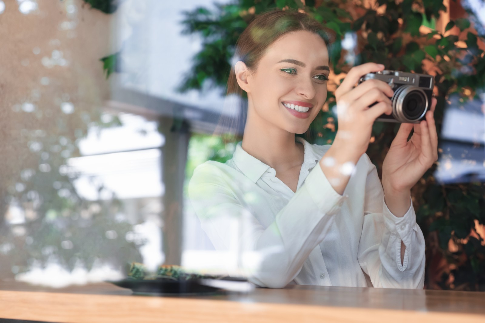 Photo of young woman with camera taking photo at cafe, view through window. Creative hobby