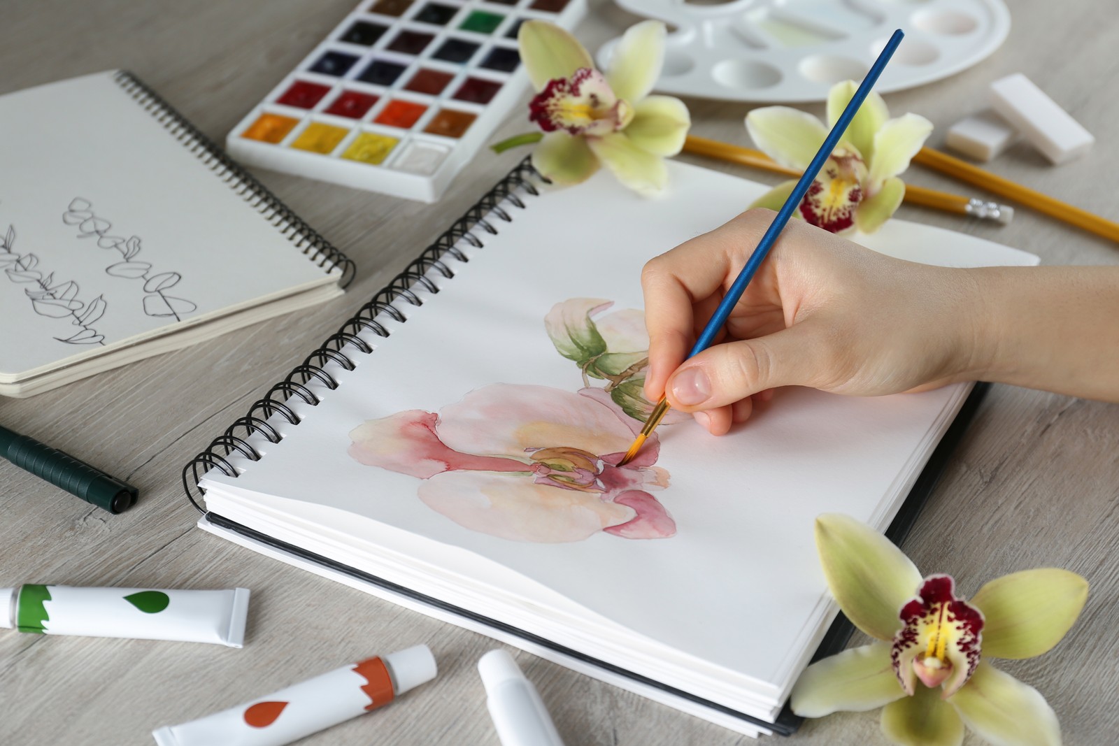 Photo of woman drawing beautiful orchid flowers in sketchbook at wooden table, closeup