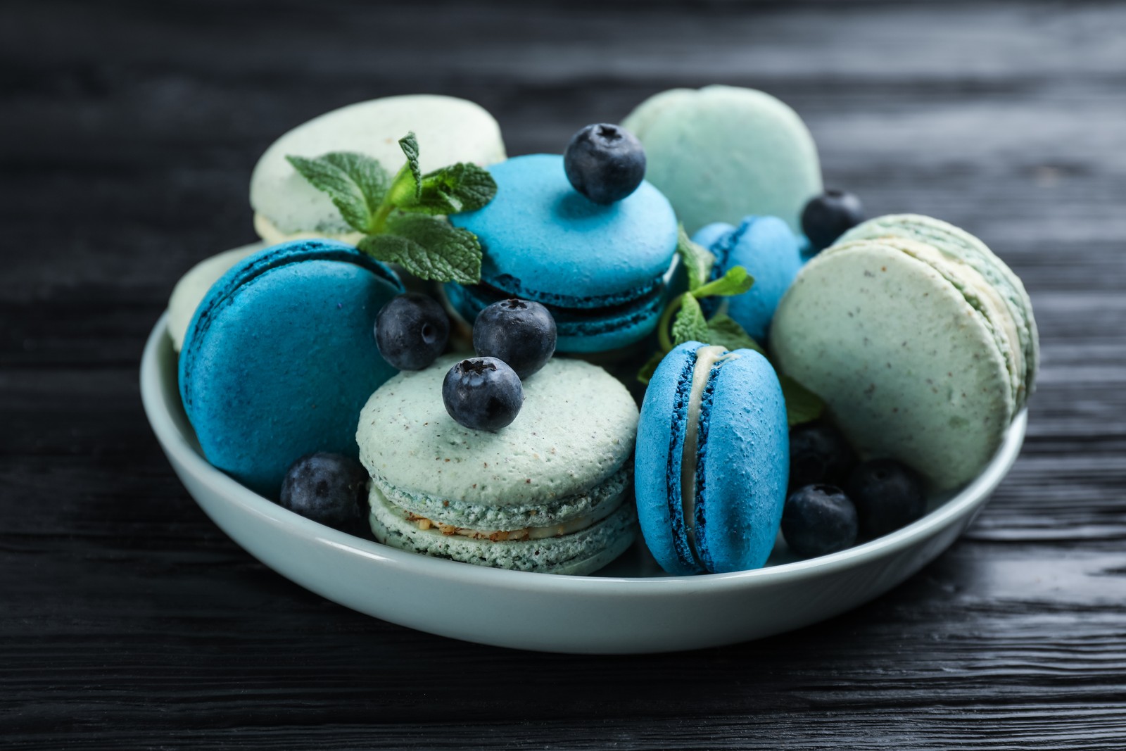 Photo of delicious macarons, mint and blueberries in bowl on black wooden table, closeup