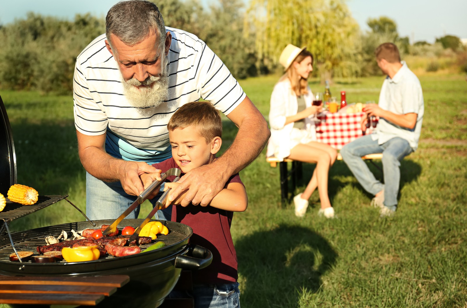 Photo of grandfather with little boy cooking food on barbecue grill and their family in park