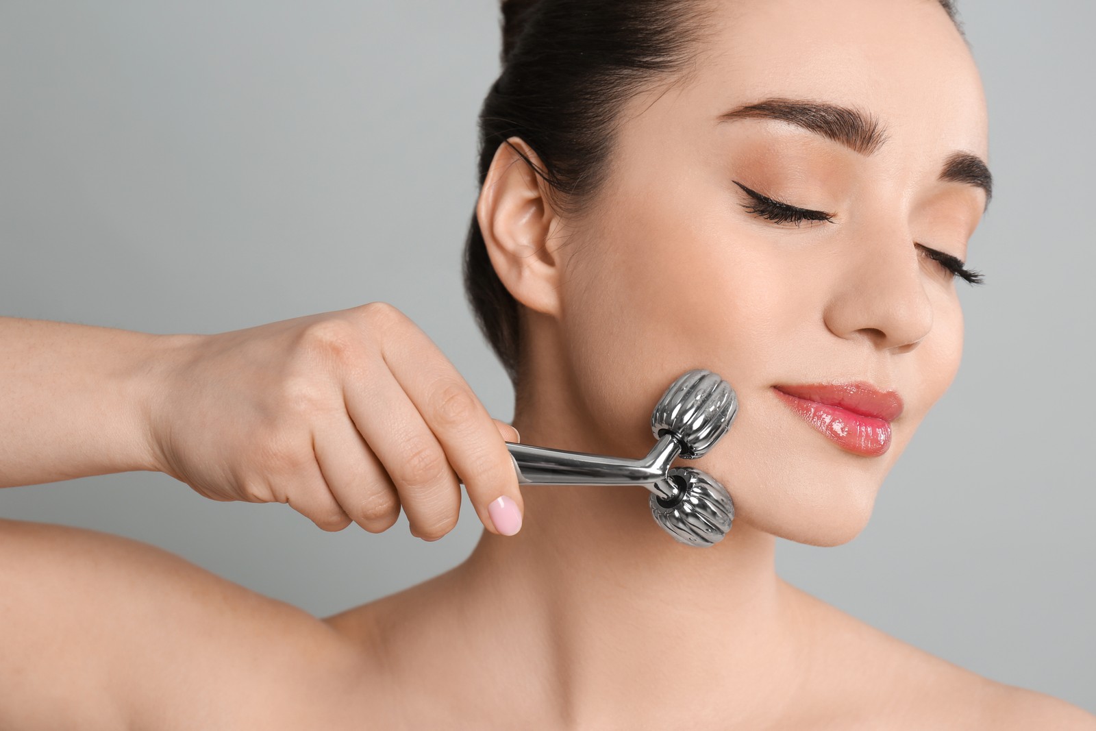 Photo of woman using metal face roller on grey background, closeup