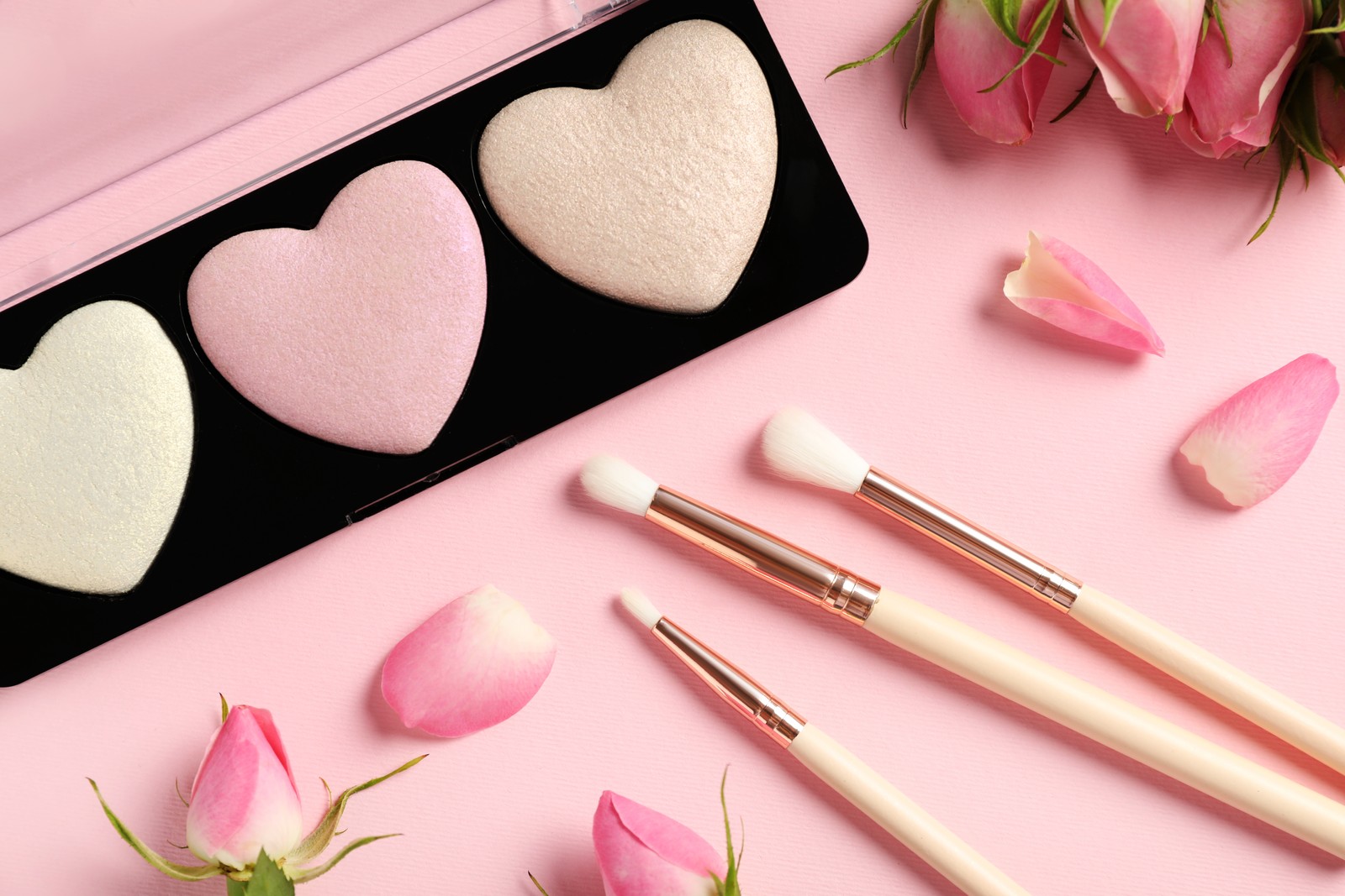 Photo of palette of heart shaped eyeshadows with brushes and roses on light pink background, flat lay