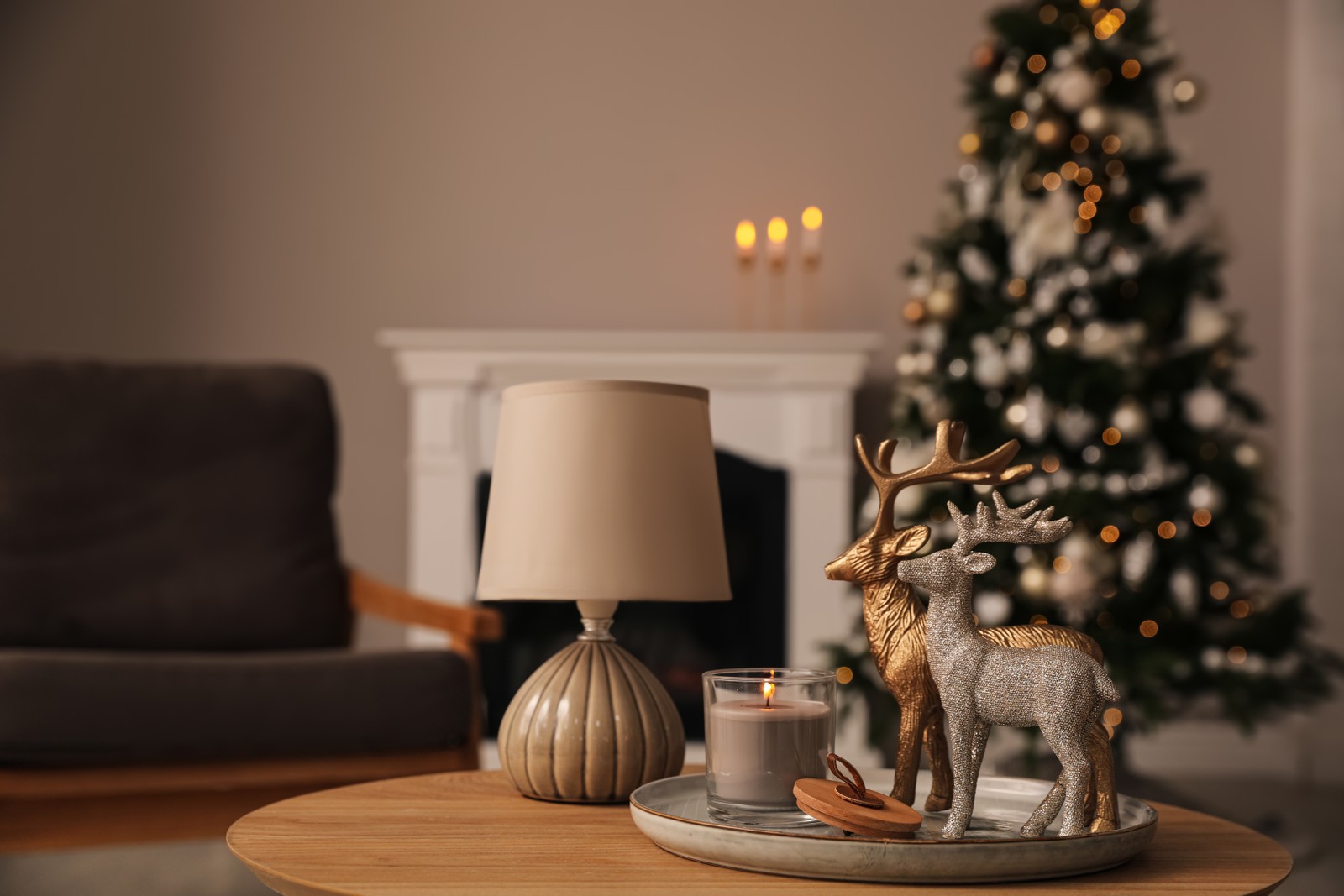 Photo of deer figures, burning candle with lamp on wooden table and Christmas tree in living room