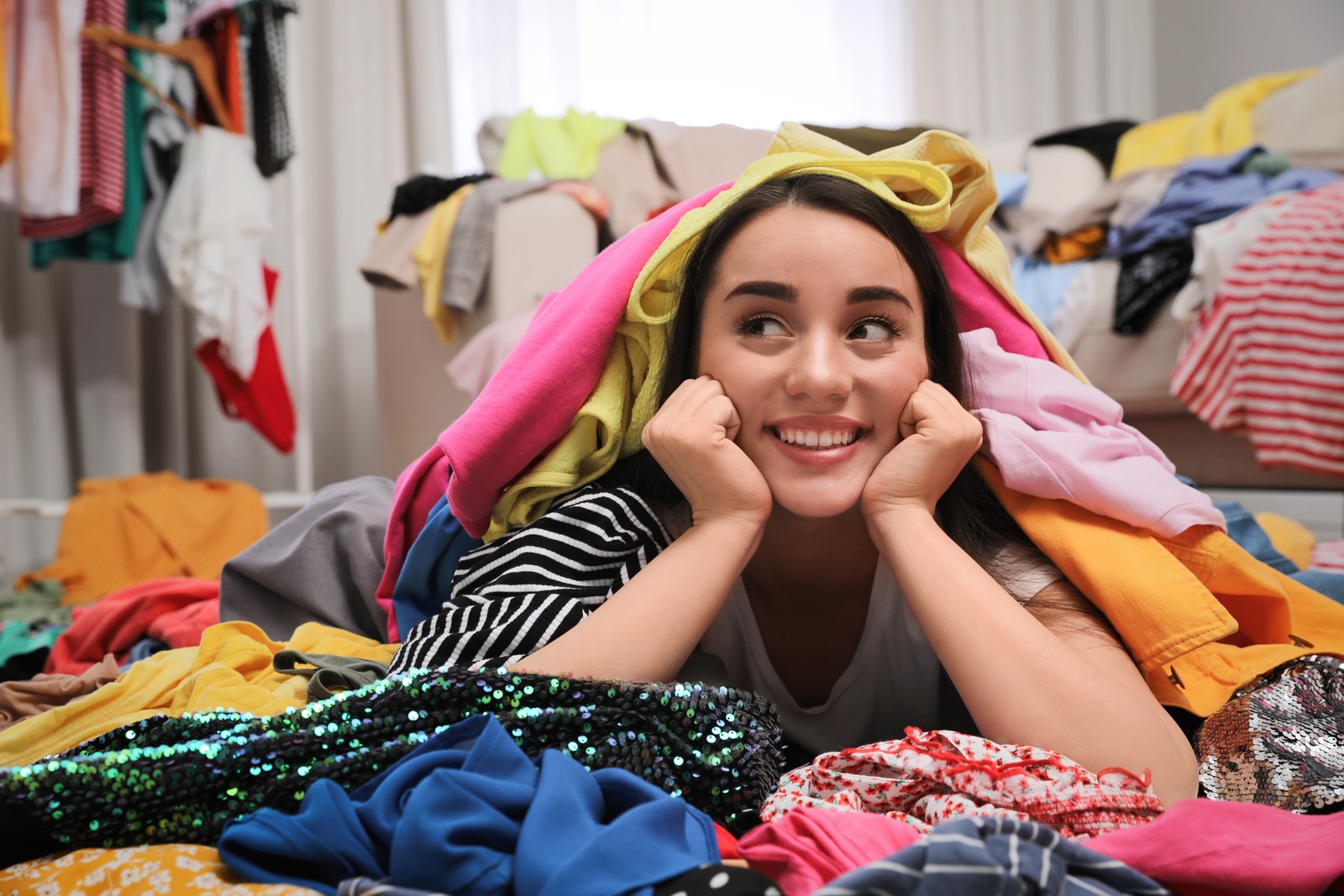 Photo of happy young woman with lots of clothes on floor in room. Fast fashion
