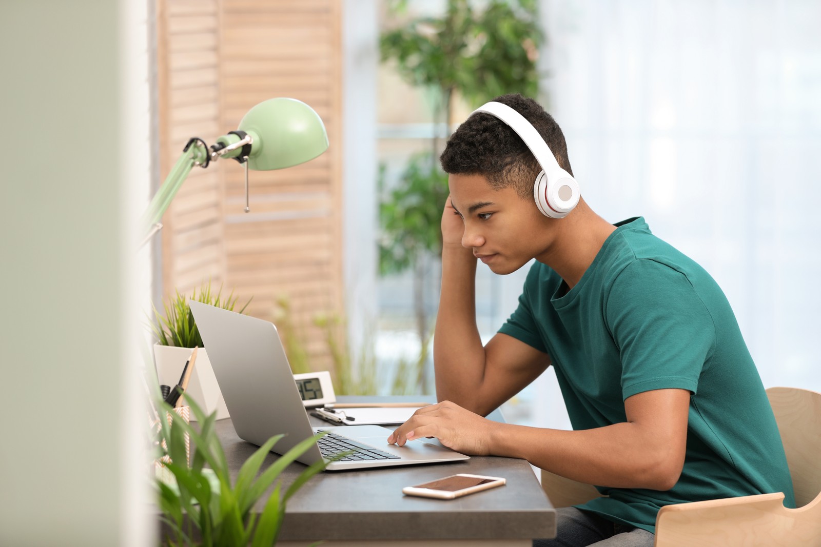 Photo of African-American teenage boy with headphones using laptop at table in room
