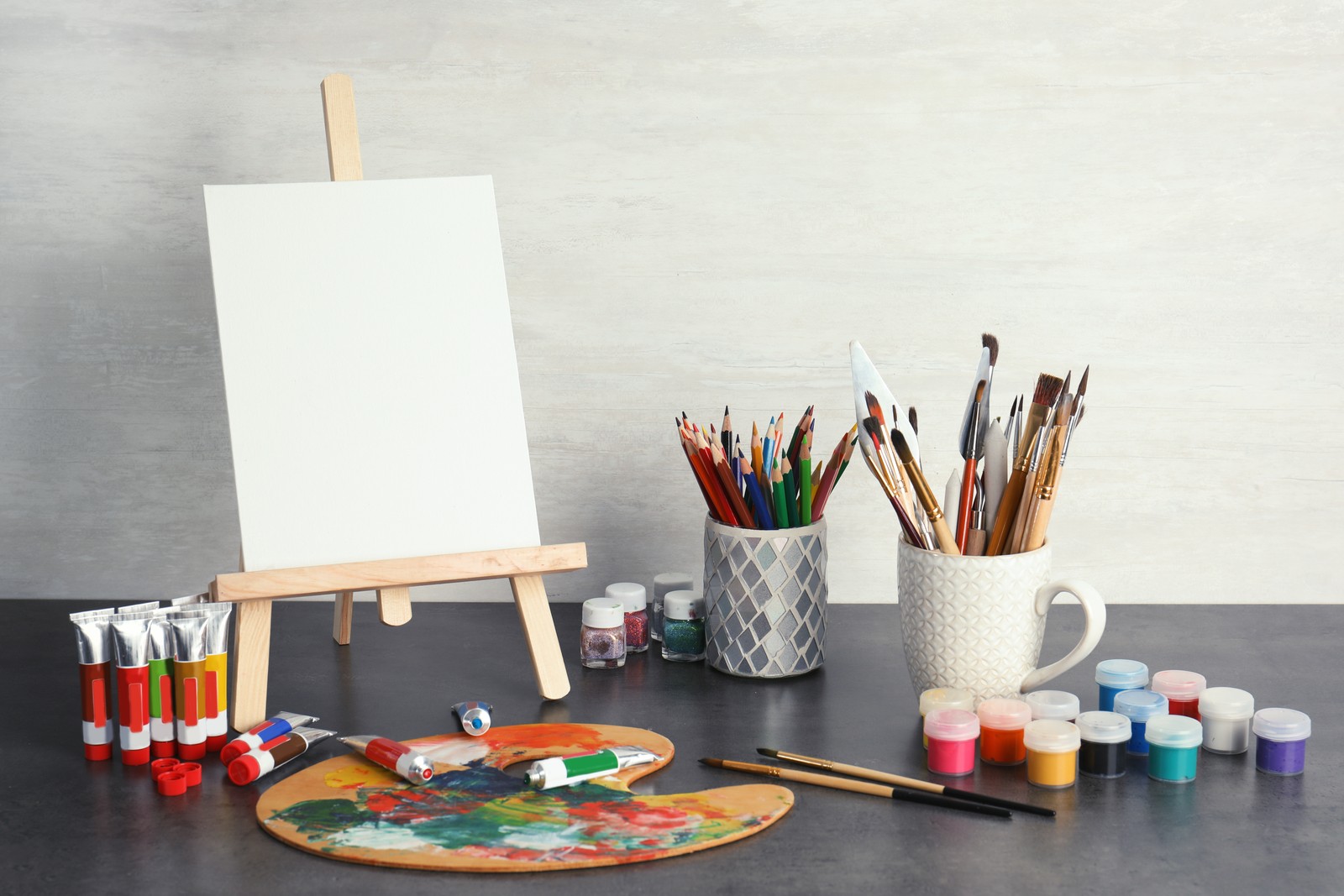 Photo of easel with various artist tools on table against light wall. Space for text