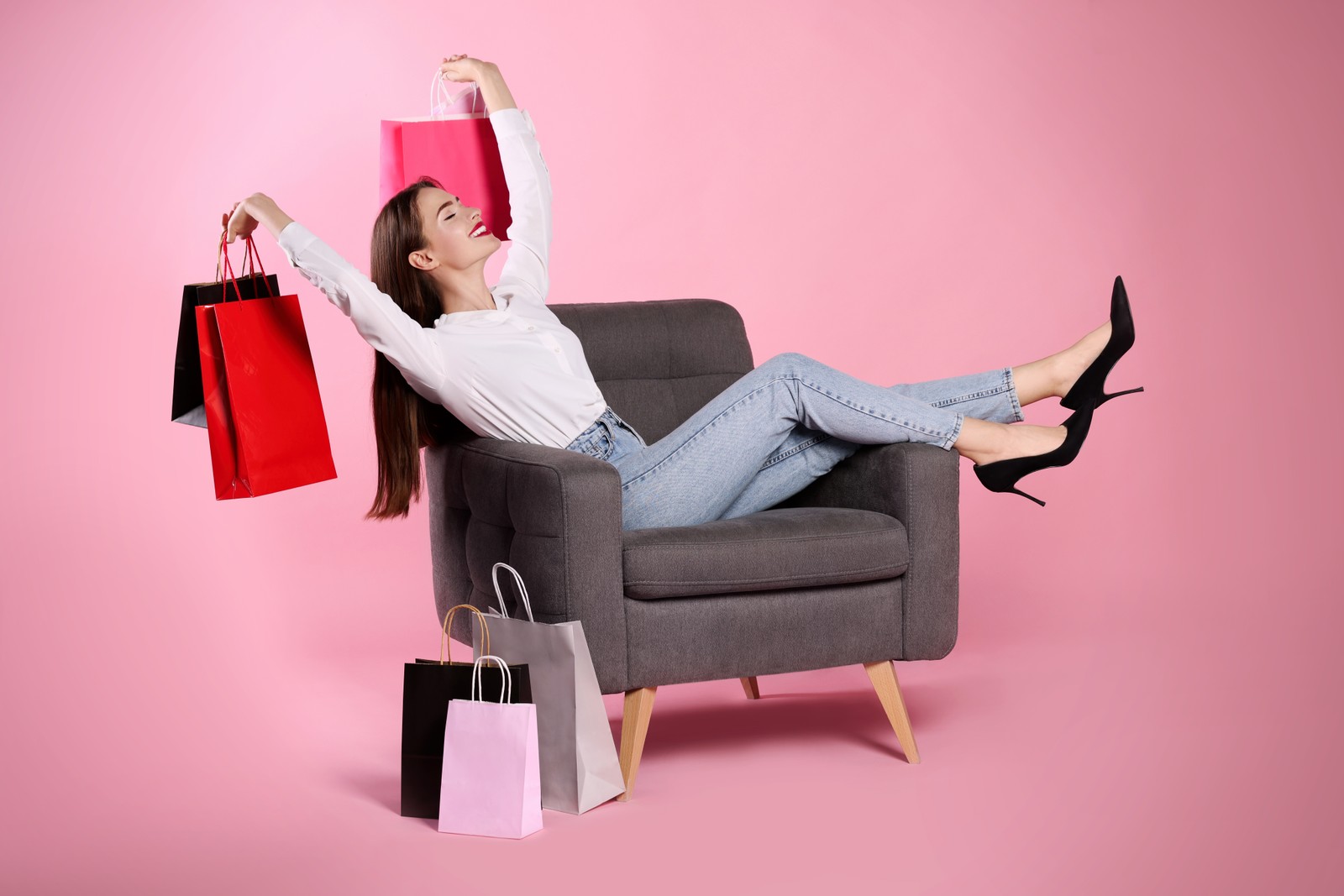 Photo of happy young woman with shopping bags on armchair against light pink background. Big sale